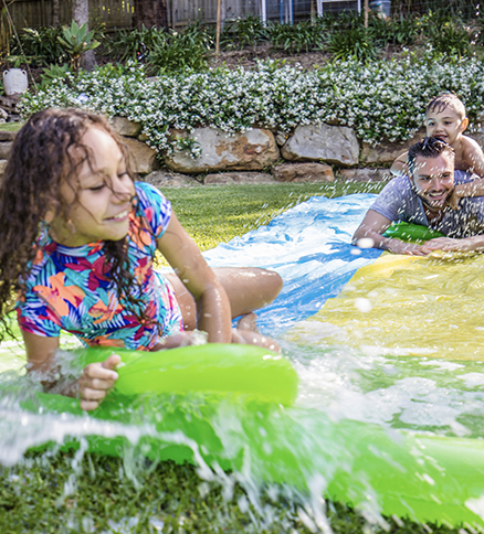 Young child sliding down a Slip-N-Slide toward the end. Behind her, her father is sliding as well, with a young child hanging onto his back.