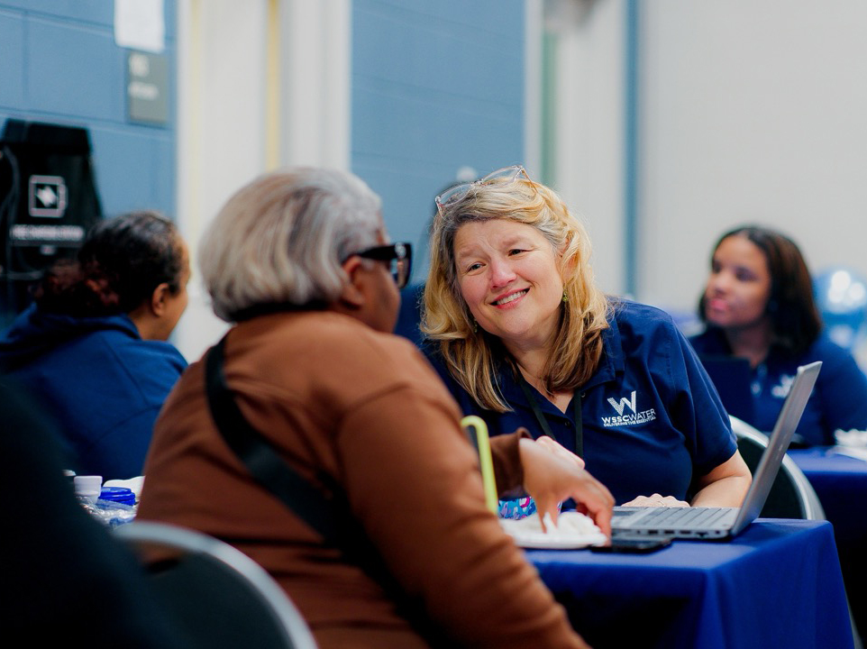 WSSC Water customer service adviser meeting with a customer at a public outreach event. They sit together at a table with a laptop between them. The WSSC Water CSA is smiling at the customer.