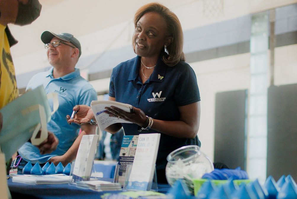 Two WSSC Water employees working a table at a public outreach event. One stands in the background, smiling at someone out of camera focus. He is wearhing a dark blue WSSC Water hat and a light blue WSSC Water polo shirt. The other employee in the foreground is speaking to a customer. She is wearing a dark blue WSSC Water polo and holds a notepad and pen.
