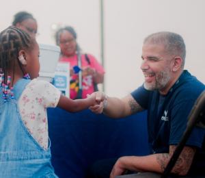 WSSC Water employee kneeling to the level of a little girl shaking hands with him. He is wearing a dark blue WSSC Water T-shirt and smiling brightly at the young girl.