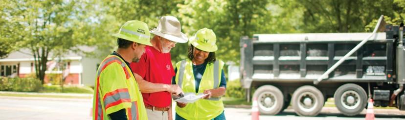 Group of three WSSC Water employees, wearing appropriate safety gear including hardhats and reflective vests, study a document one of them holds. A WSSC Water truck and orange traffic cones are in the background. 