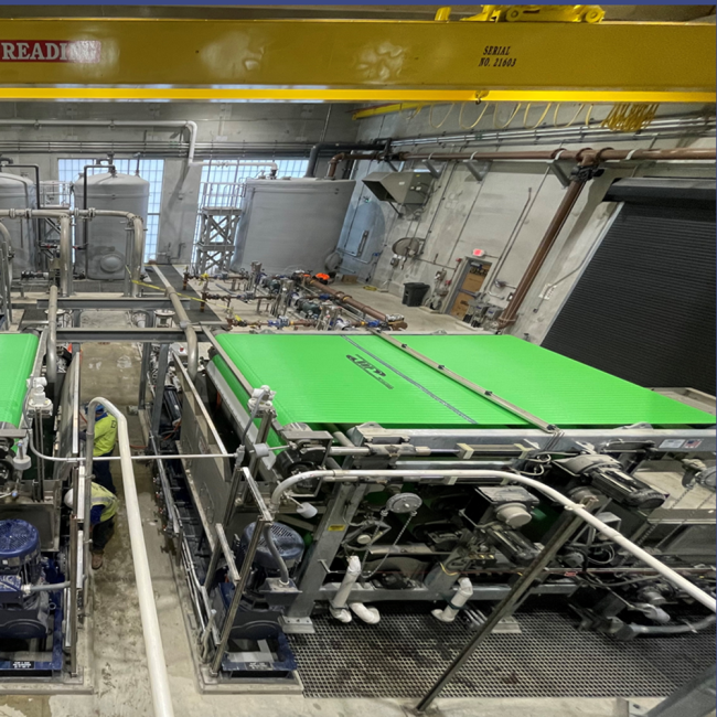 Photo of the belt filter presses taken from the mezzanine level in the Solids Building.  Workers are fine tuning and calibrating the controls.