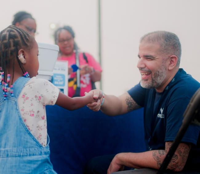 WSSC Water employee kneeling to the level of a little girl shaking hands with him. He is wearing a dark blue WSSC Water T-shirt and smiling brightly at the young girl.